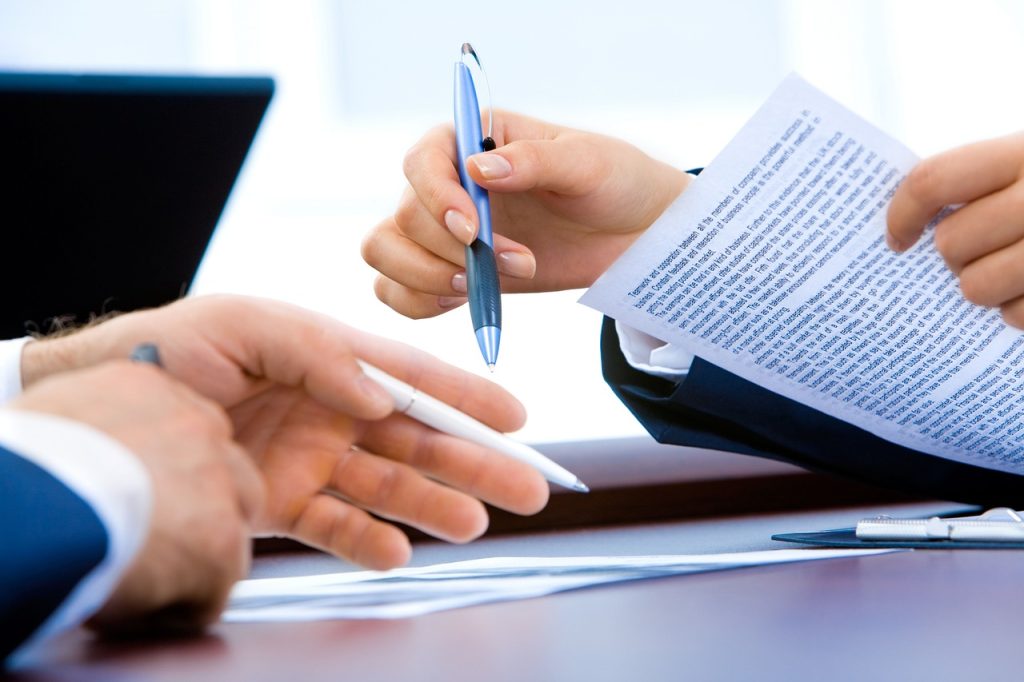 A hand holding a pen over a document on a desk beside a laptop and notebook.