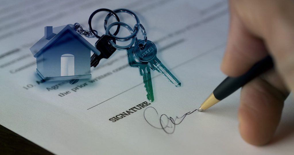 A hand signing a document next to a small model house, representing a real estate sale or property purchase.