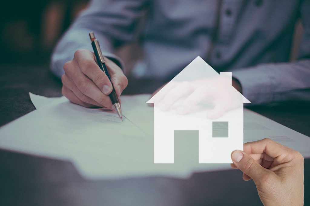 A hand holds a pen over a paper contract beside a model house, representing a home purchase or mortgage agreement.